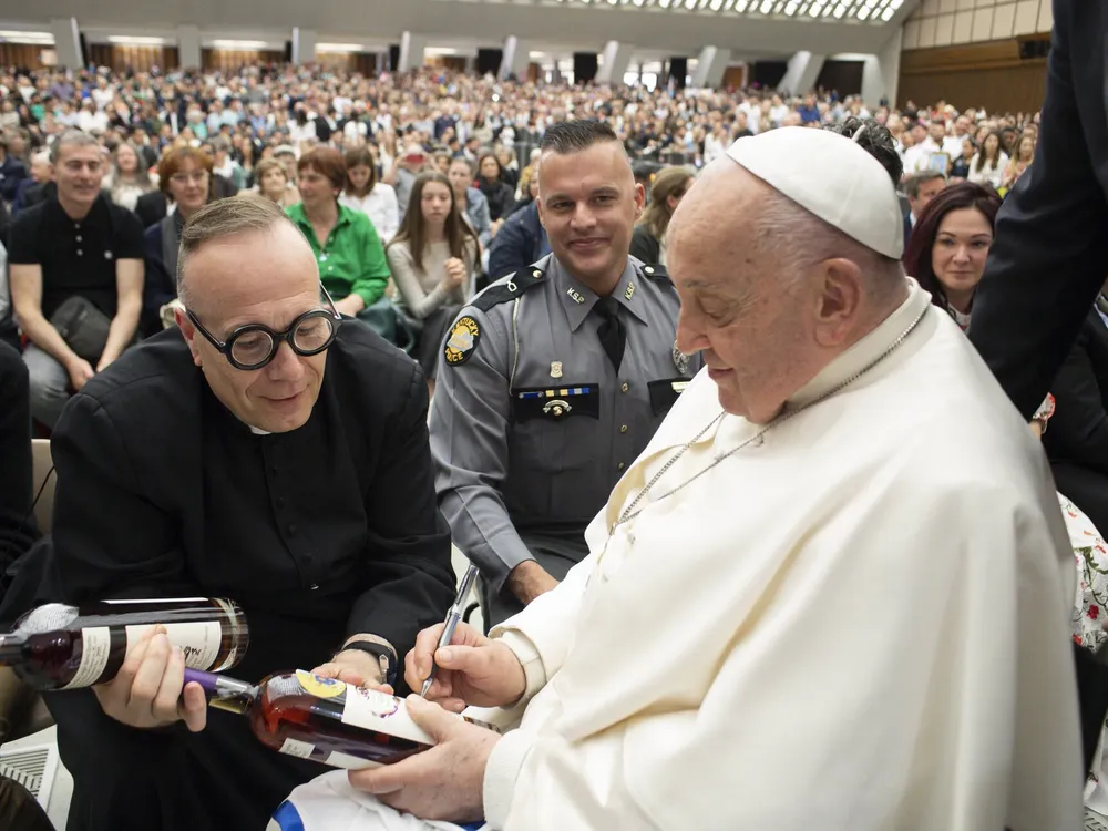 A man wearing glasses holding a bottle next to a man wearing a white robe and a little white hat signing a bottle