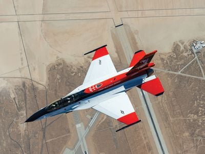 A sleek jet aircraft painted red, white, and black flies over an air base in the desert.