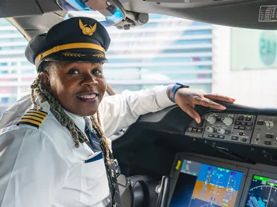 A 65-year-old Black woman sits in the left seat of an airliner cockpit. She is smiling and wearing an airline captain's uniform, including a white shirt and a hat.