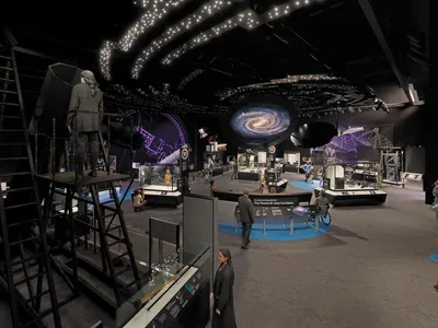 An artist's renderign of a cavernous museum exhibit space with a dark ceiling illuminated by images of bright white stars and a spiral galaxy. On the floor of the gallery, numerous visitors wander past glass display cases filled with artifacts.