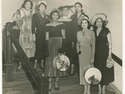 A black-and-white photograph of seven women standing on a flight of stairs. Mae Reeves stands to the farthest right on the lowest step and wears a white hat and floral patterned handbag.