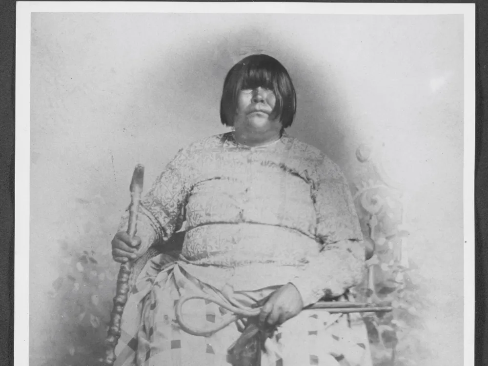 Black and white portrait of Dat So La Lee seated in a chair and surrounded by six decorative baskets on the floor by her feet.