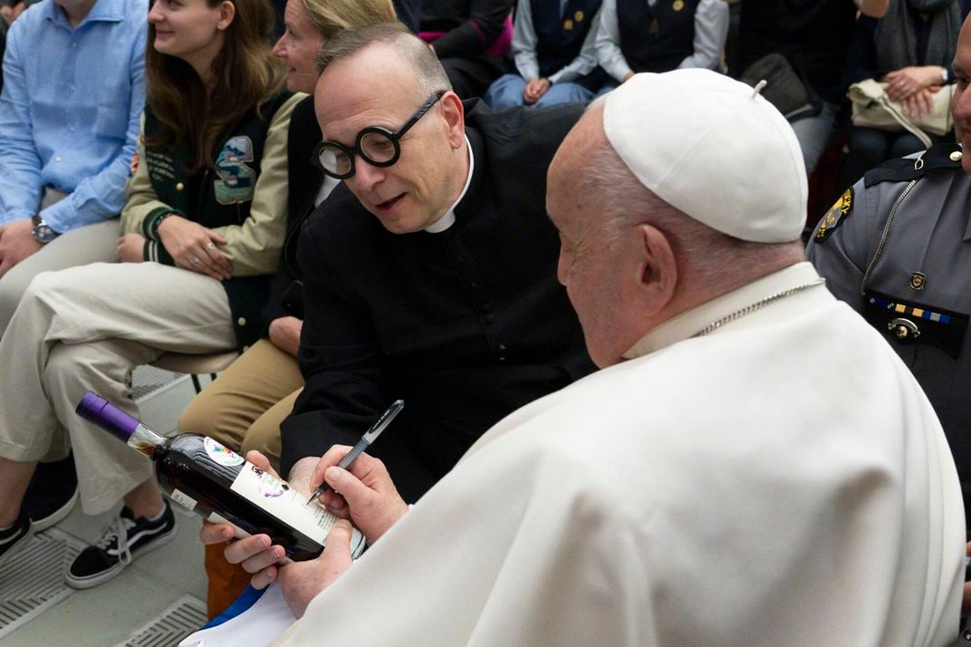 Man wearing a little white hat and a white robe signing a bottle next a man in a black robe with black glasses