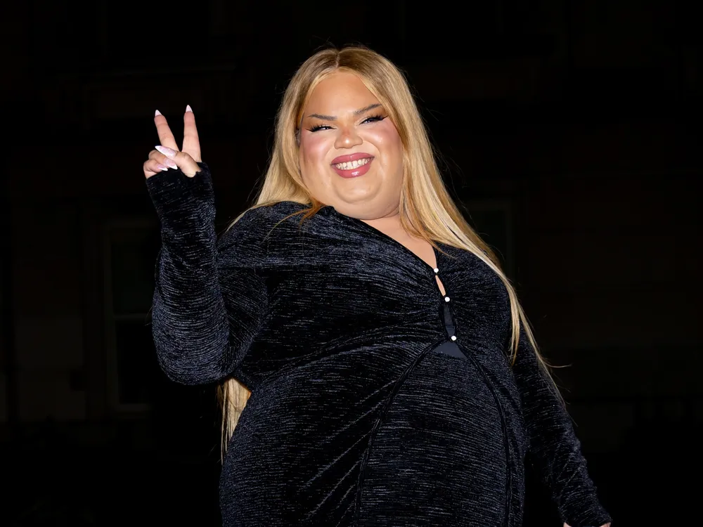 Woman with blond hair wearing black and holding up one hand with a peace sign