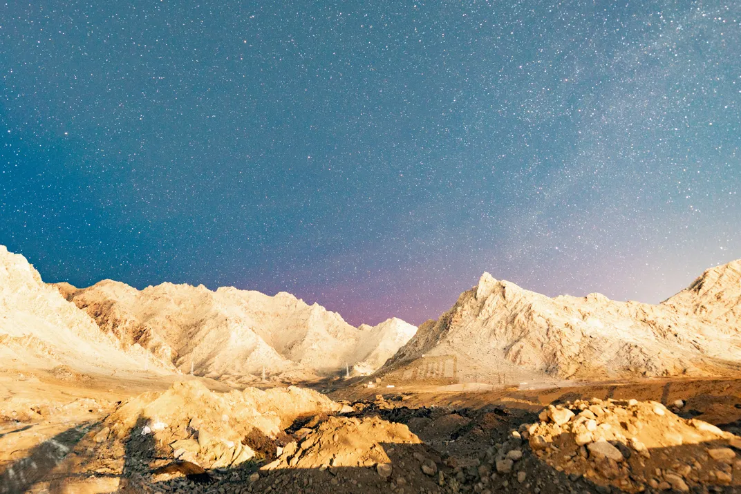 a starry sky appears bright over dry, vegetation-less hills, with a faint glow of red over the top of the peaks