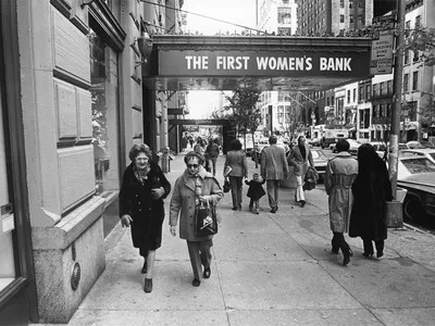 Candid black and white photo of people walking down the street. Two women face the camera in the foreground under an awning with the words “The First Women’s Bank” on it.